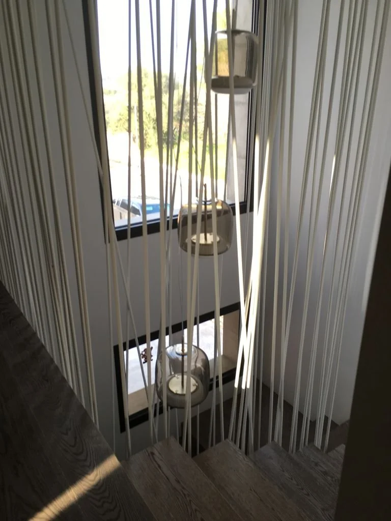 Escalera de madera con barandales de cuerdas y lámparas colgantes, vista desde la parte superior hacia abajo, con ventana y paisaje exterior visible.