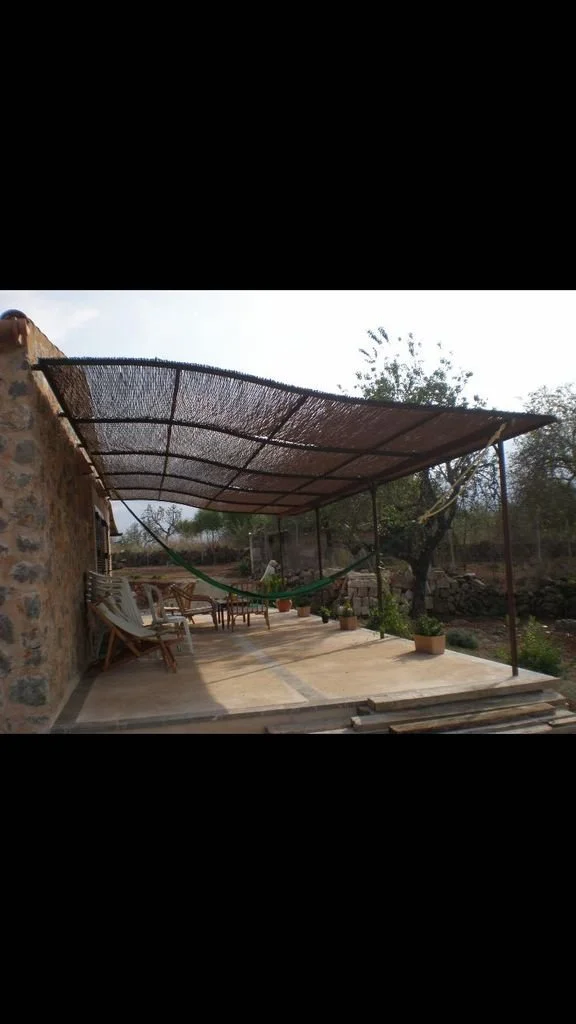 Área exterior con sombra de estructura tejada, muebles de madera, sombrilla y plantas enmacetadas, en un ambiente rural con paisaje natural.