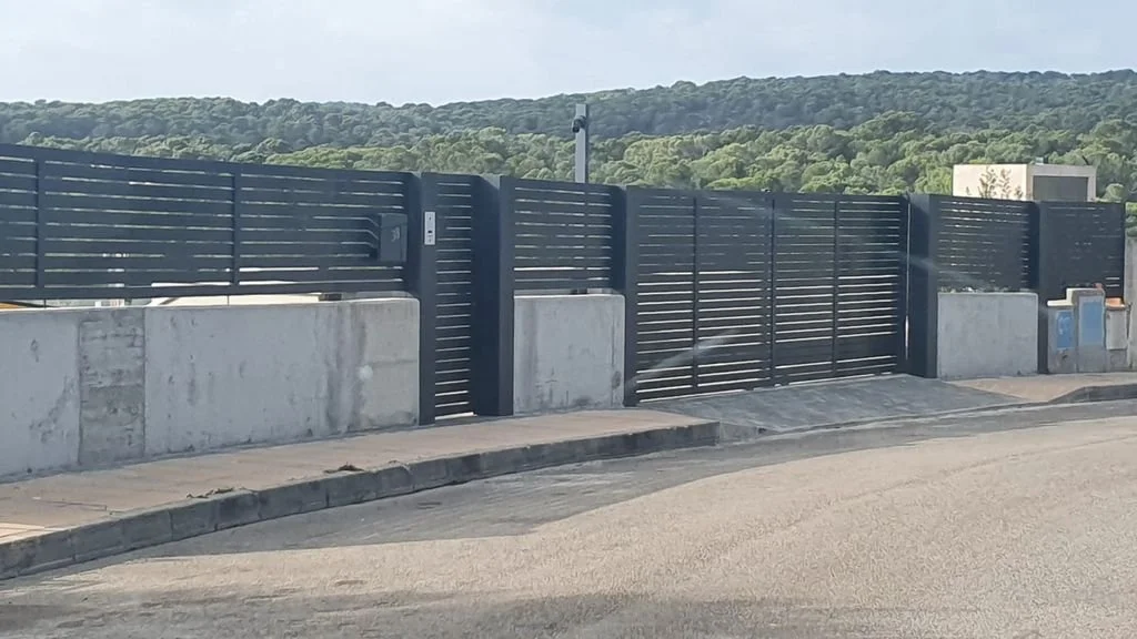 Puerta metálica negra con muro de concreto y cercado de tablas horizontales, en una calle con acera y colina verde en el fondo.