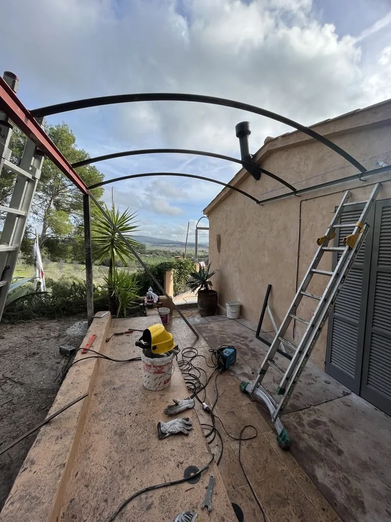Área de construcción con herramientas, escaleras, un casco amarillo, y plantas en el fondo, en un porche con cielo nublado.