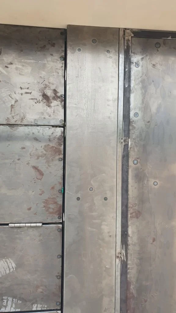 Puerta o panel de metal con tornillos visibles y marcas de uso o suciedad, instalado en una pared o estructura.