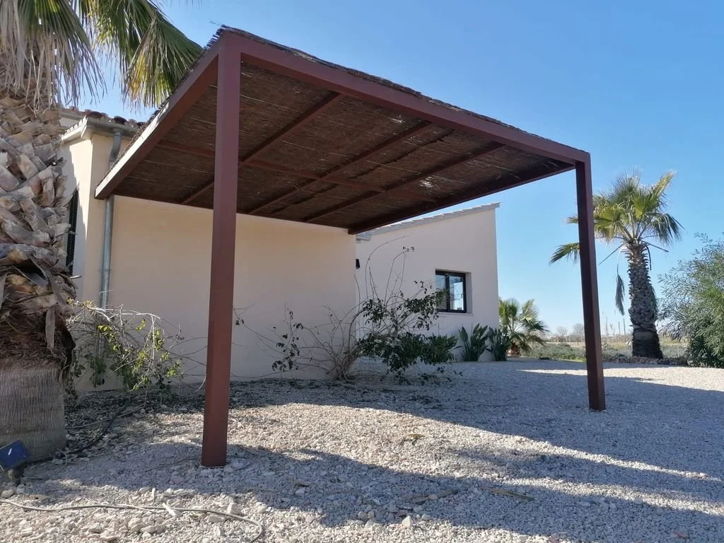 Estructura de una pérgola de madera en el exterior de una casa blanca, con plantas y palmeras en un clima árido y soleado.
