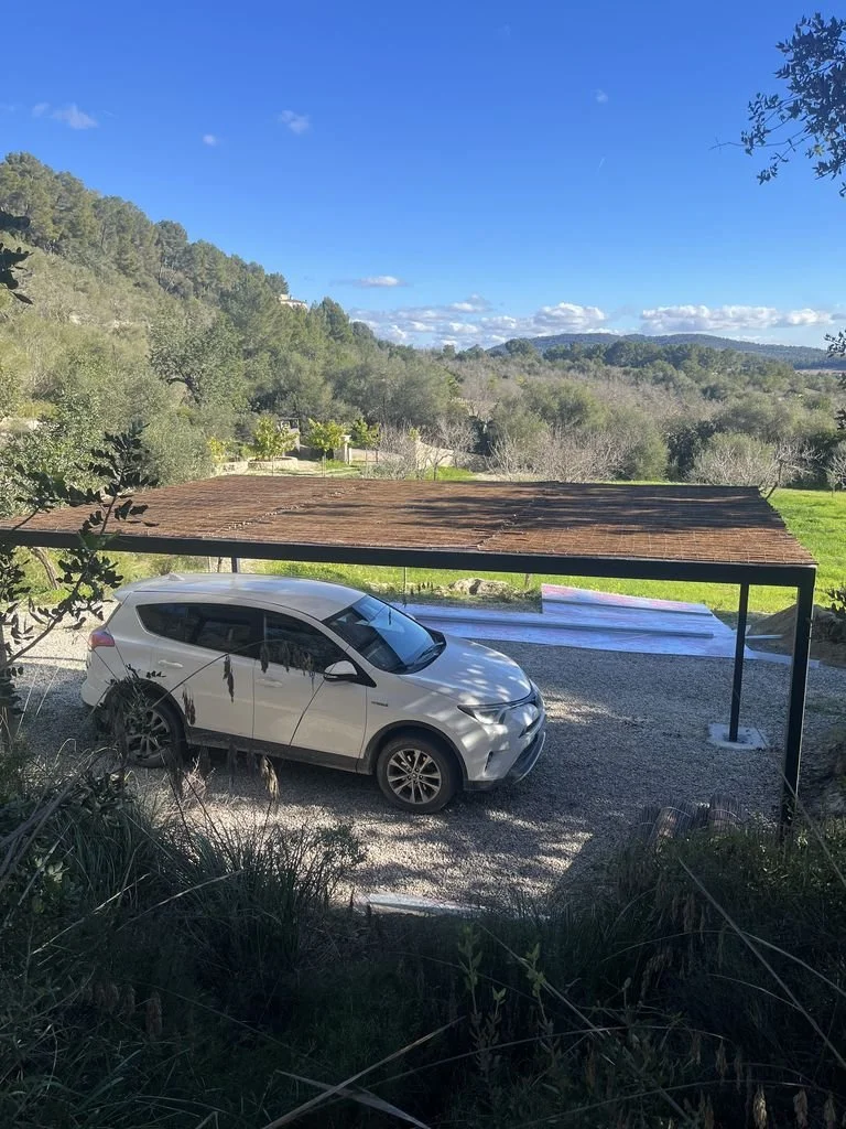 Cochera techada con techo de madera y columnas negras, con un coche plateado estacionado sobre suelo de grava, rodeada de naturaleza con árboles y colinas de fondo bajo cielo azul con algunas nubes.