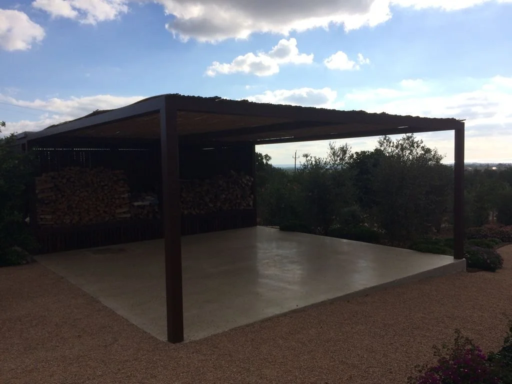 Estructura moderna al aire libre con techo de madera, piso de concreto y leña apilada al fondo, rodeada de naturaleza y árboles.
