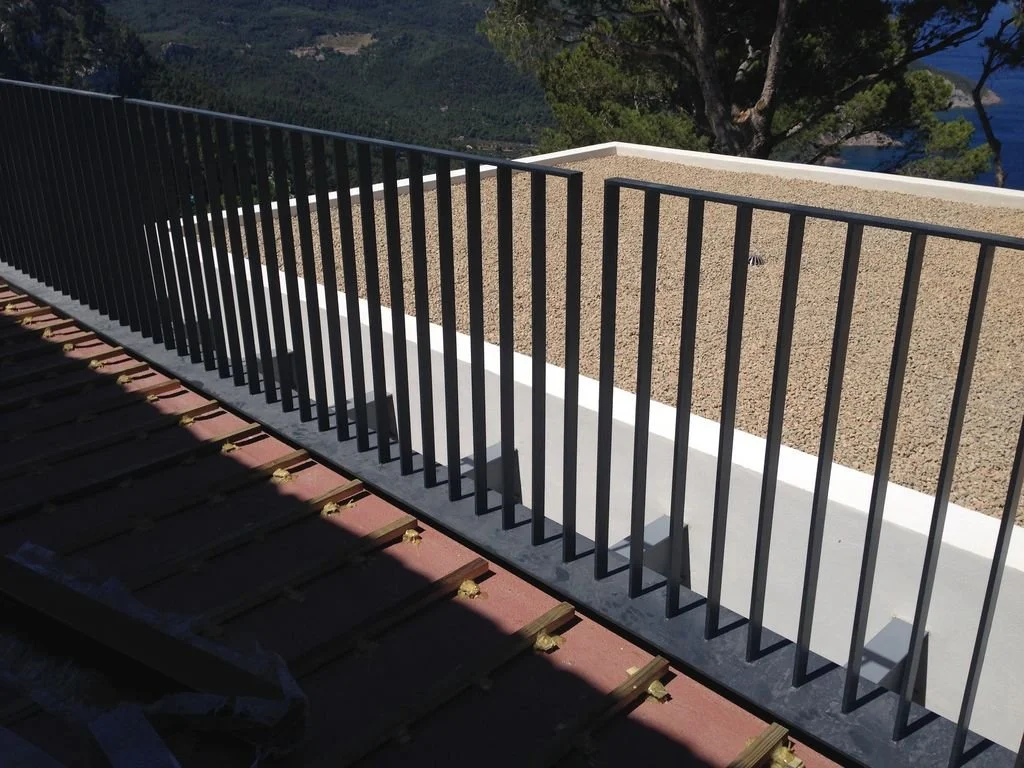 Vista desde una terraza o balcón con barandilla de barras negras y superficie de grava, con árboles y paisaje montañoso al fondo.