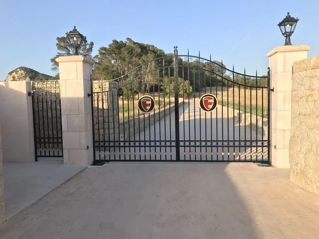 Puerta de entrada de hierro con dos pilares de piedra y faroles, que da acceso a un camino rodeado de árboles y campos