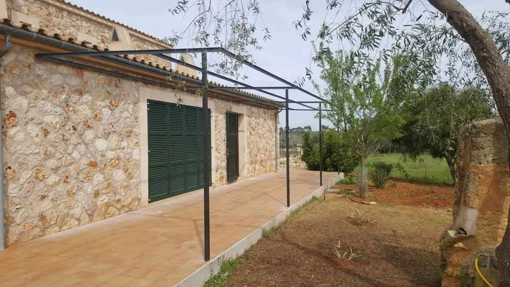 Casa de piedra con patio y estructura metálica para sombra, en un entorno rural con árboles y césped.