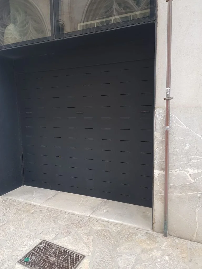 Puerta de garaje negra con diseño lineal, ubicada en la entrada de un edificio, con revestimientos de piedra y una tubería de metal a la derecha.
