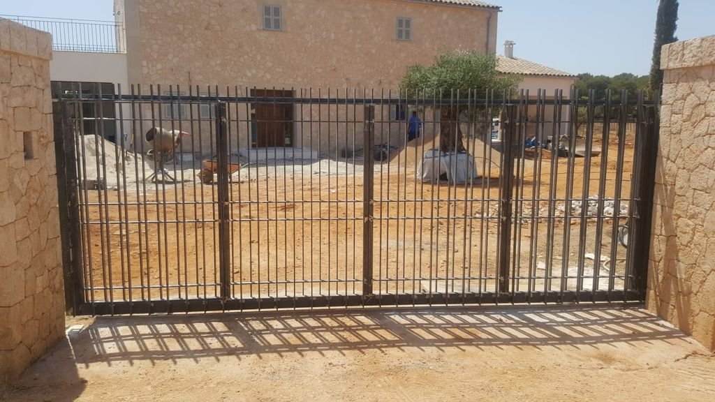 Puerta negra de barrotes que da hacia un patio con tierra, un árbol y algunas construcciones en construcción o en proceso de terminación.