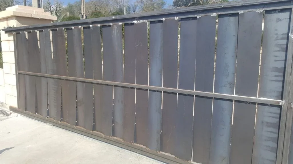 Una verja de metal grande instalada en la entrada de un terreno, con estructura de barras verticales y horizontales, en un entorno exterior con paredes y vegetación al fondo.