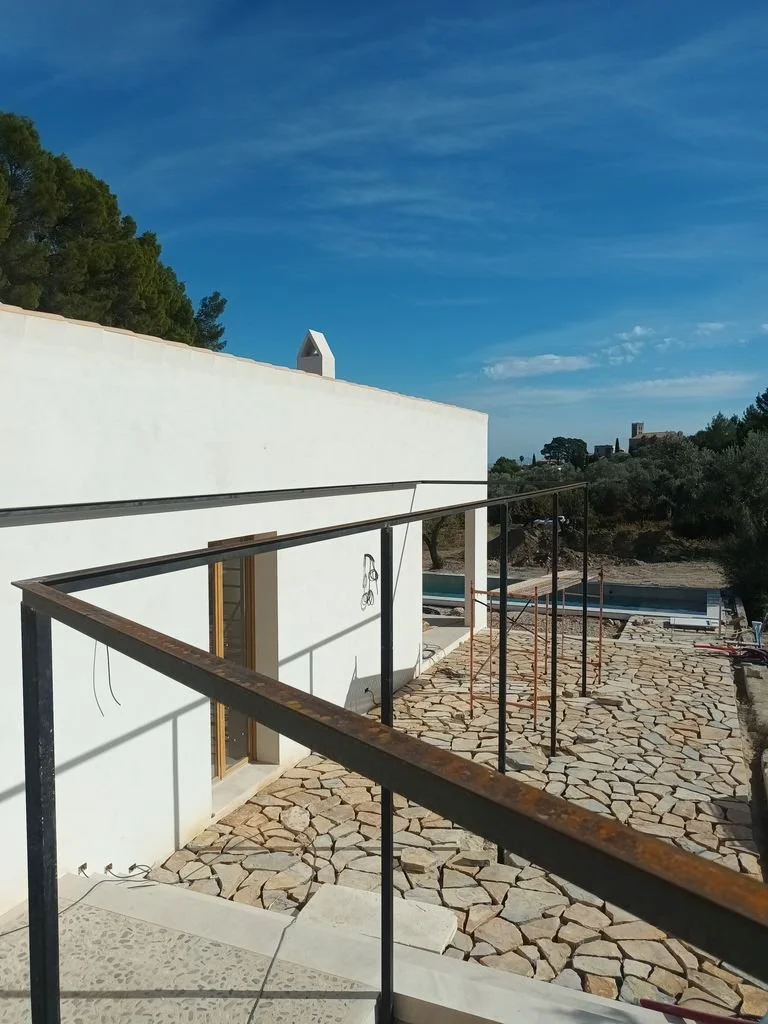 Construcción en progreso en una terraza con suelo de piedra, vista de un edificio blanco y el cielo azul con algunas nubes.