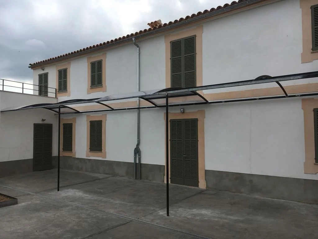 Edificio de color blanco con ventanas de persianas verdes y un toldo negro en un patio asfaltado, con cielo nublado.