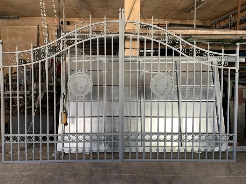 Reja de herrería blanca con detalles curvos en la parte superior y un escudo en el centro de cada puerta, ubicada en un taller o bodega.