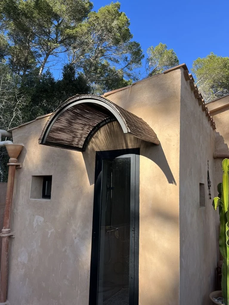 Vista de una casa con paredes de color beige, un arco de cobertura y una puerta de cristal negro, rodeada de árboles y plantas, en un día soleado con cielo azul.