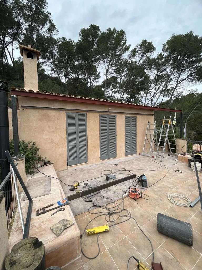 Área de patio en construcción con herramientas, escaleras, cableado y una casa con paredes de estuco y ventanas con persianas grises, rodeada de árboles.
