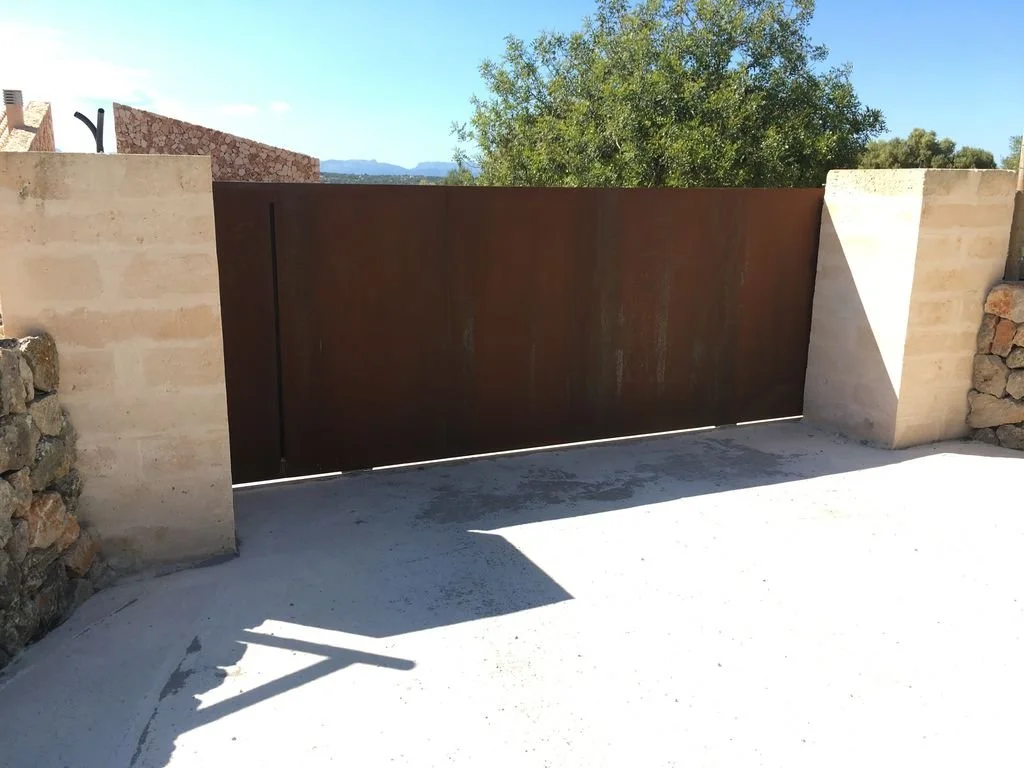Puerta metálica de color marrón en un muro, en un entorno exterior con cielo claro y árboles en el fondo, y sombra en el suelo.