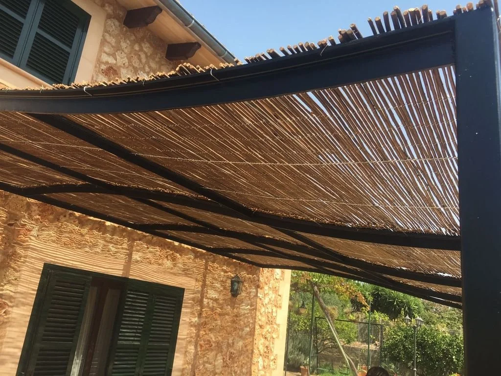 Persiana de madera en una estructura metálica en un patio con vegetación y una casa de piedra al fondo.