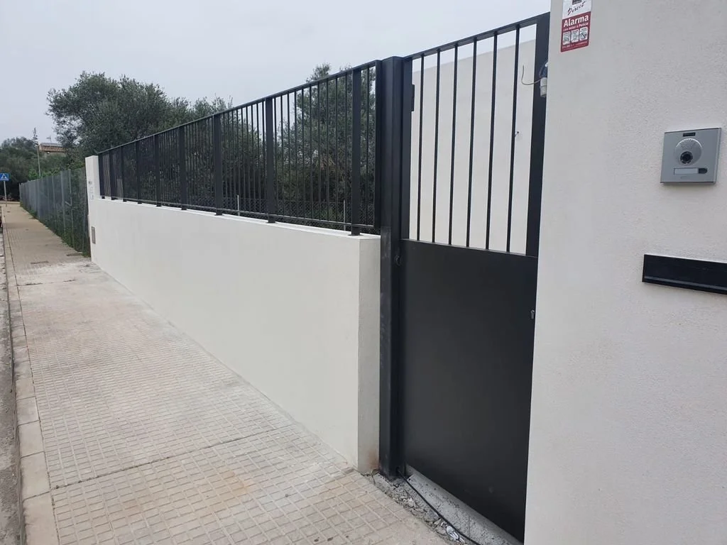 Puerta y reja negra en muro blanco junto a acera de concreto.