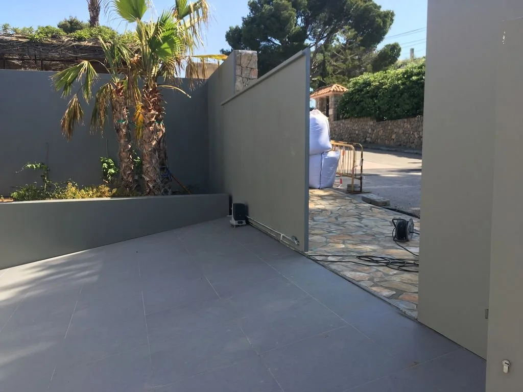 Área exterior con paredes grises, palmera, plantas en una jardinera y suelo de cerámica, en proceso de construcción o reparación con cables y equipos eléctricos.