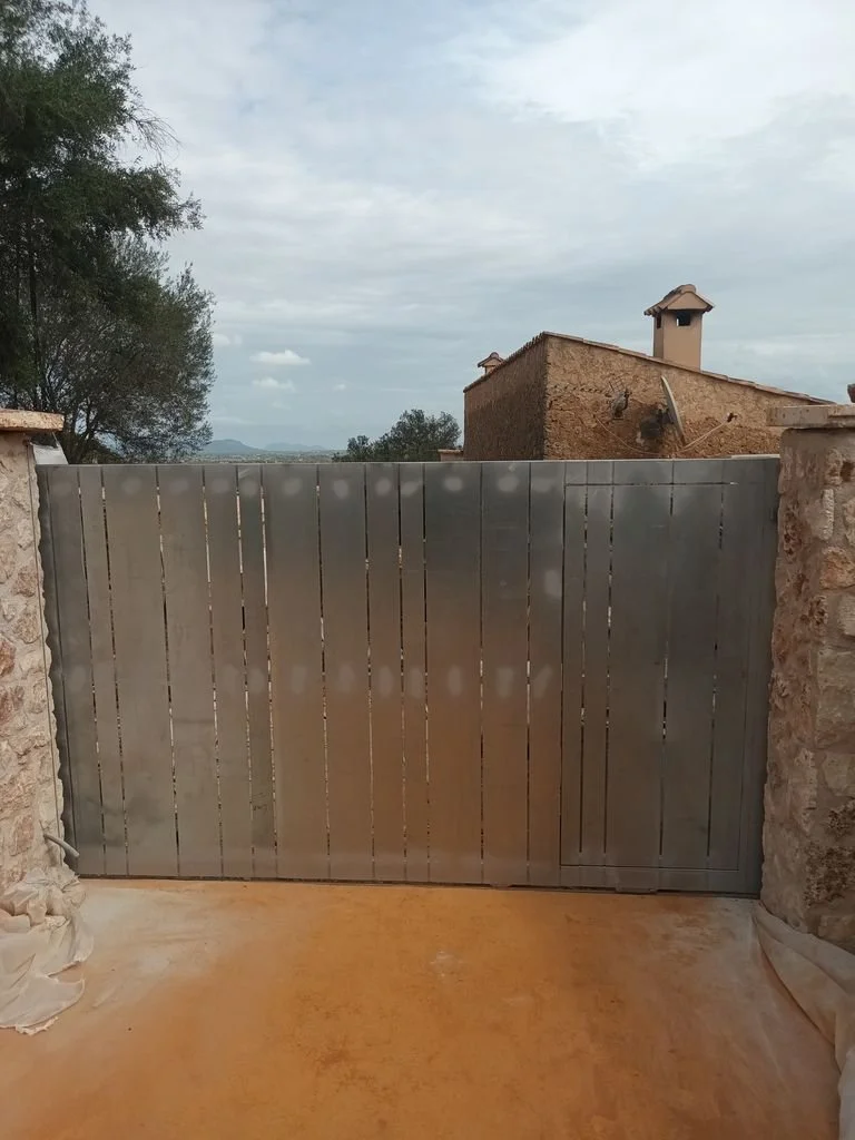 Puerta de metal en un muro de piedra, con cielo nublado y casas de barro al fondo en un entorno rural.