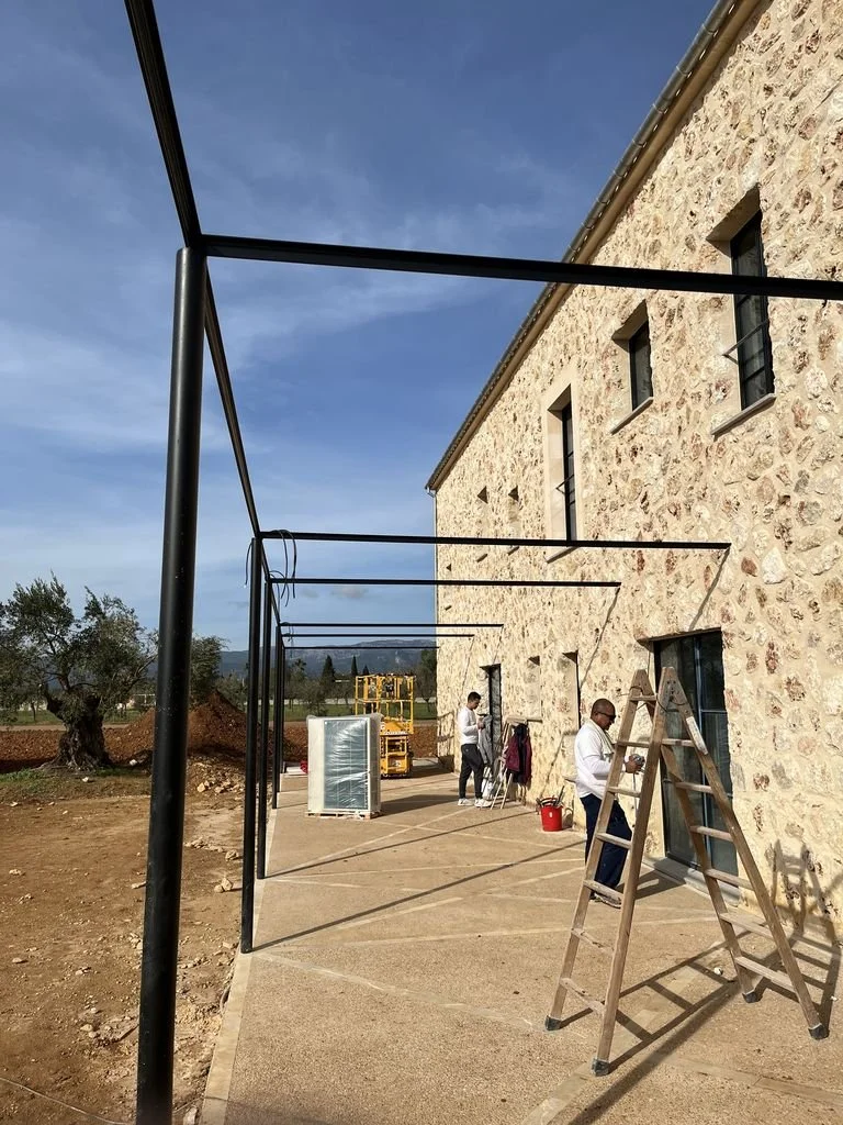 Personas instalando estructura metálica en exterior de edificio de piedra con ventanas y piso de concreto, en un día soleado.