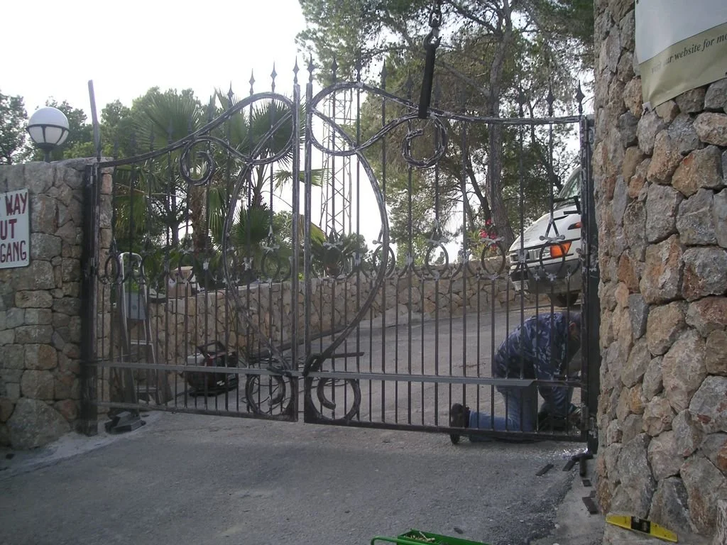 Persona arreglando una verja de hierro en una entrada rodeada de muro de piedra, con árboles y plantas en el fondo y un automóvil estacionado.
