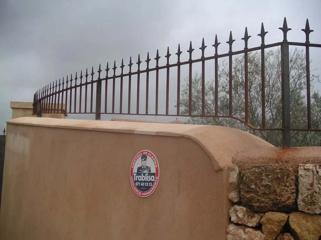 Valla de hierro con picas en la parte superior sobre un muro de piedra y concreto, con aviso de protección policial en español.