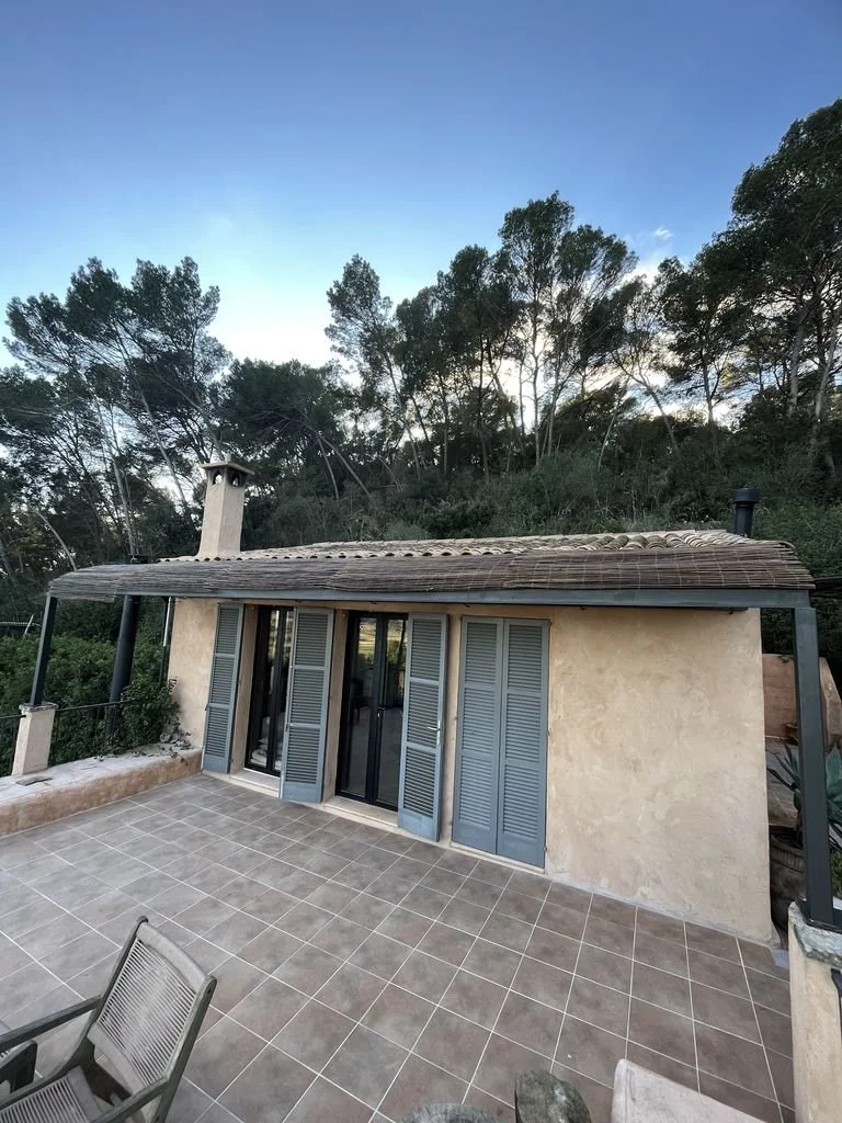 Terraza exterior con piso de cerámica, vista a un bosque con árboles altos y cielo despejado, casa de una planta con paredes de estuco y ventanas con puertas plegables de madera pintadas de azul.