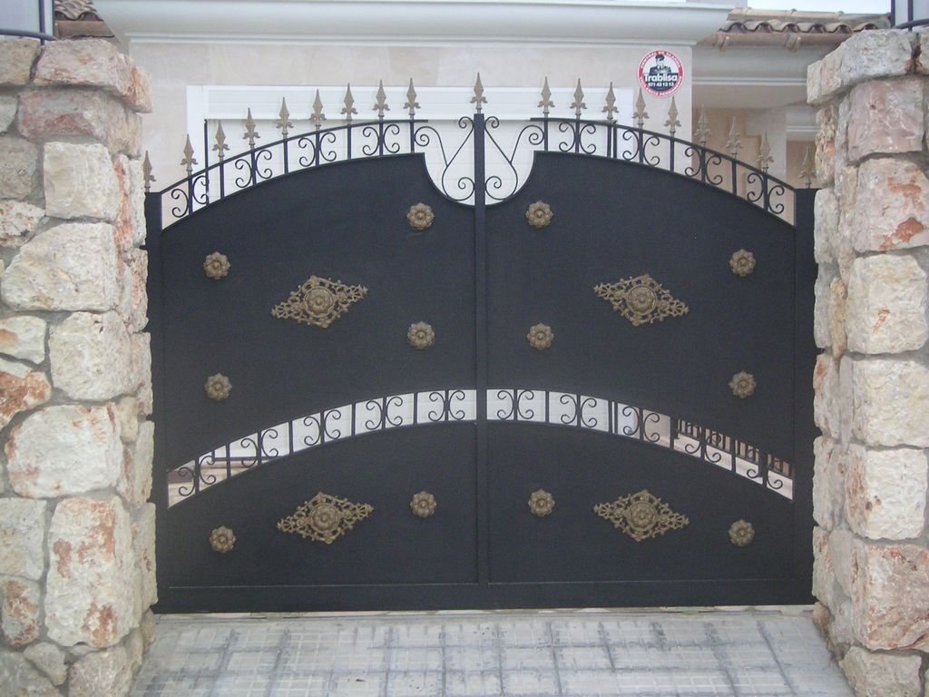 Puerta de hierro negra con detalles dorados, rodeada por muros de piedra