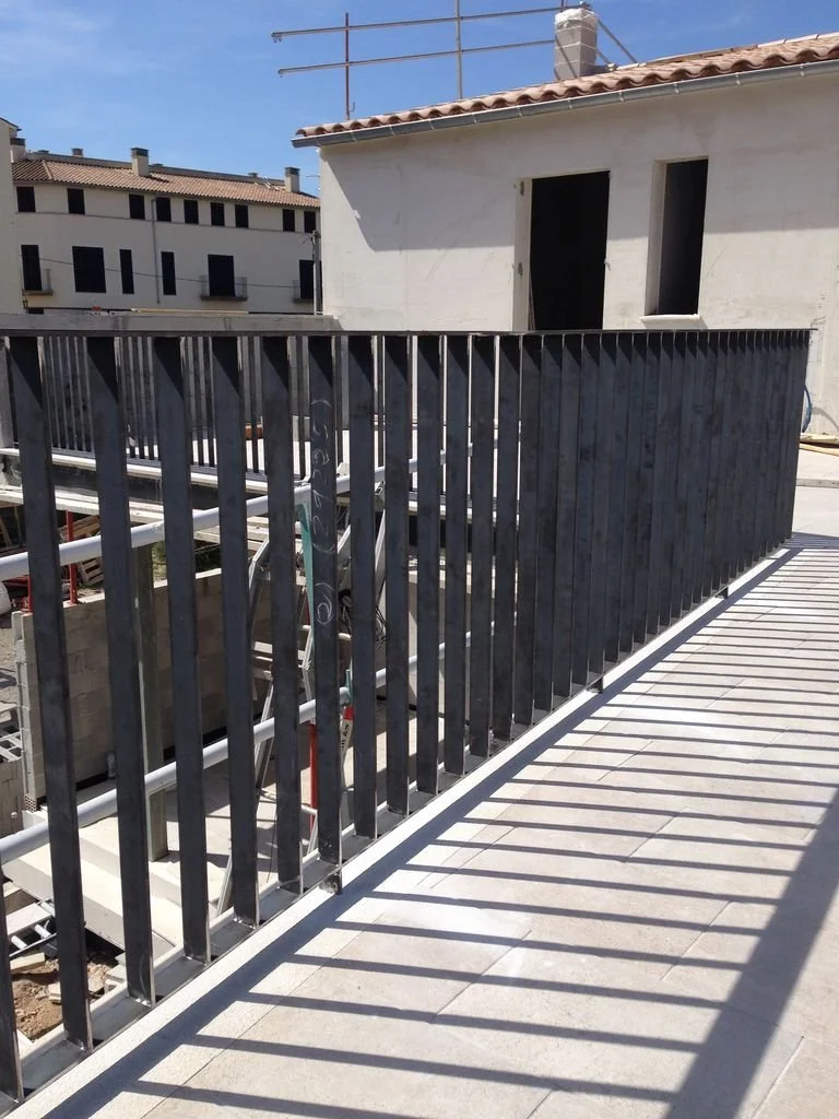 Barandal negro en una terraza sobre baldosas blancas, con sombras proyectadas por el sol y un edificio en construcción al fondo.