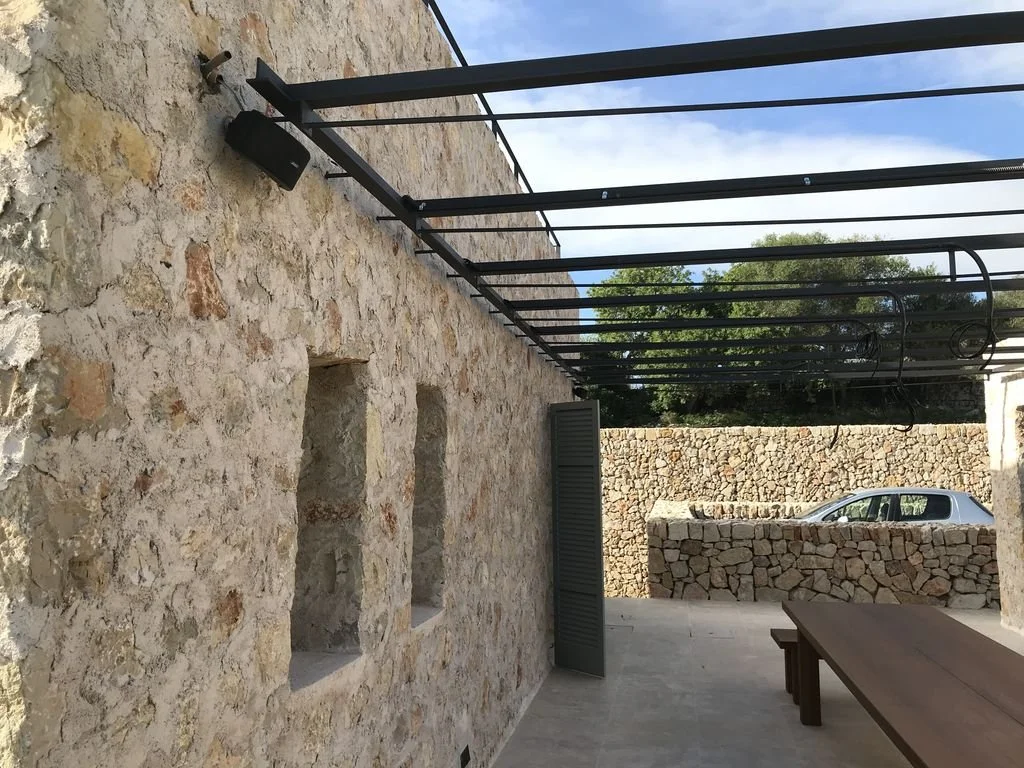 Vista de una pared de piedra con dos ventanas pequeñas, una puerta con persiana abierta, un coche gris estacionado y una estructura metálica sobre un patio con banca de madera.