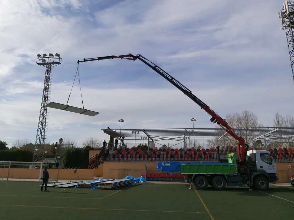 Camión de pluma colocando una estructura en un campo de fútbol, con espectadores en las gradas y torres de alumbrado en el fondo.