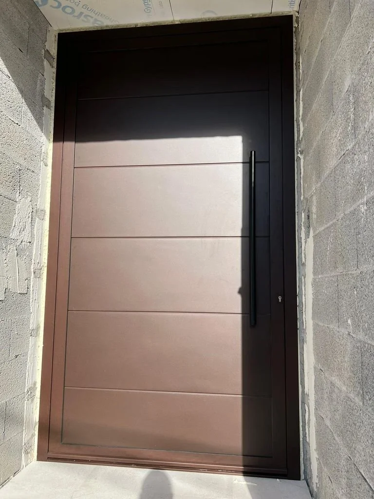 Puerta moderna de madera enmarcada por paredes de bloques de cemento, con manija larga negra y en construcción.