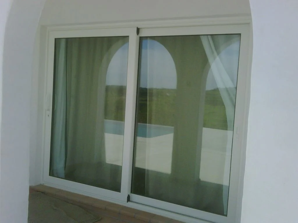 Puerta de cristal corrediza, con cortinas blancas, en un balcón con vista a un paisaje verde.