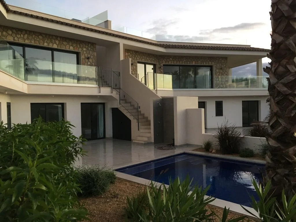 Casa moderna de dos pisos con balcón de vidrio, escaleras exteriores, piscina en el jardín y vegetación, en un entorno soleado.