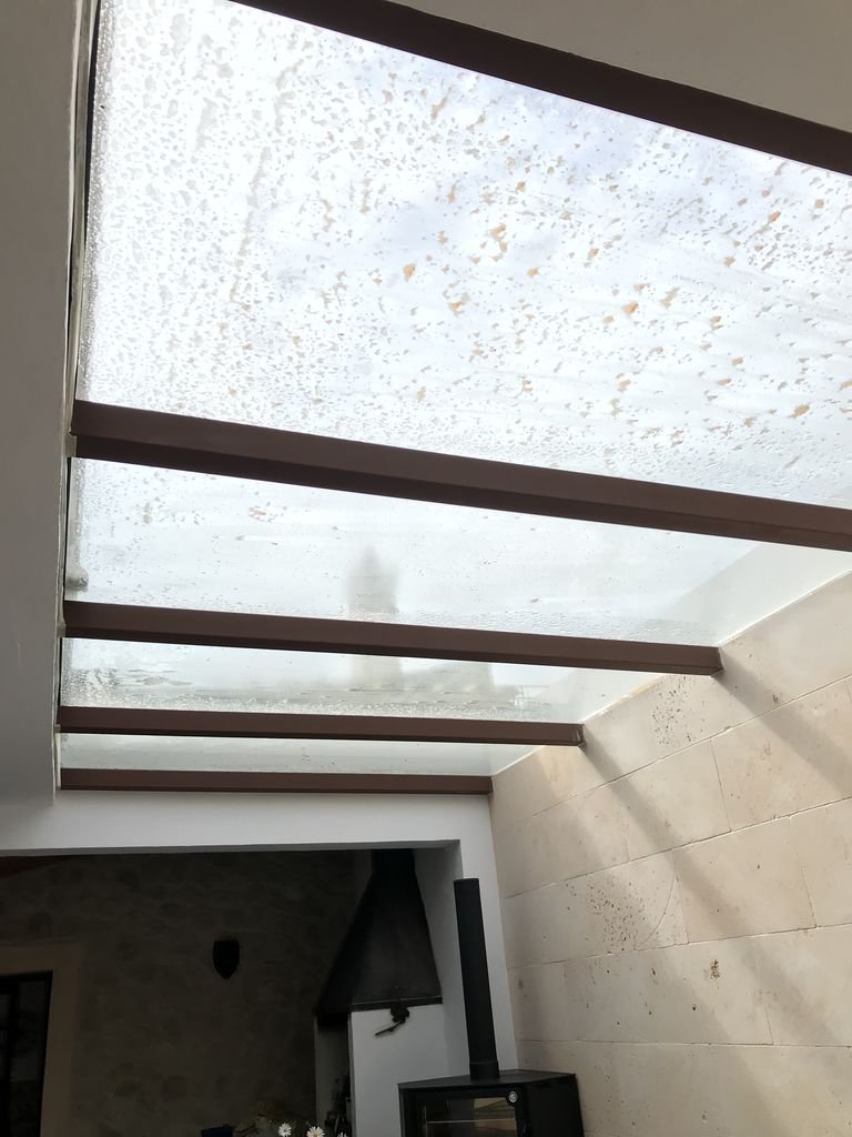  Techo de vidrio con marcas de agua y estructura de varias vigas de madera o metal, en un espacio interior con pared de piedra y chimenea.