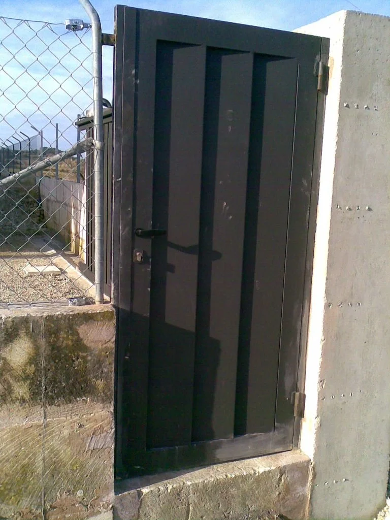 Puerta metálica negra en una cerca de alambre con un muro de concreto a un lado y cielo despejado arriba.