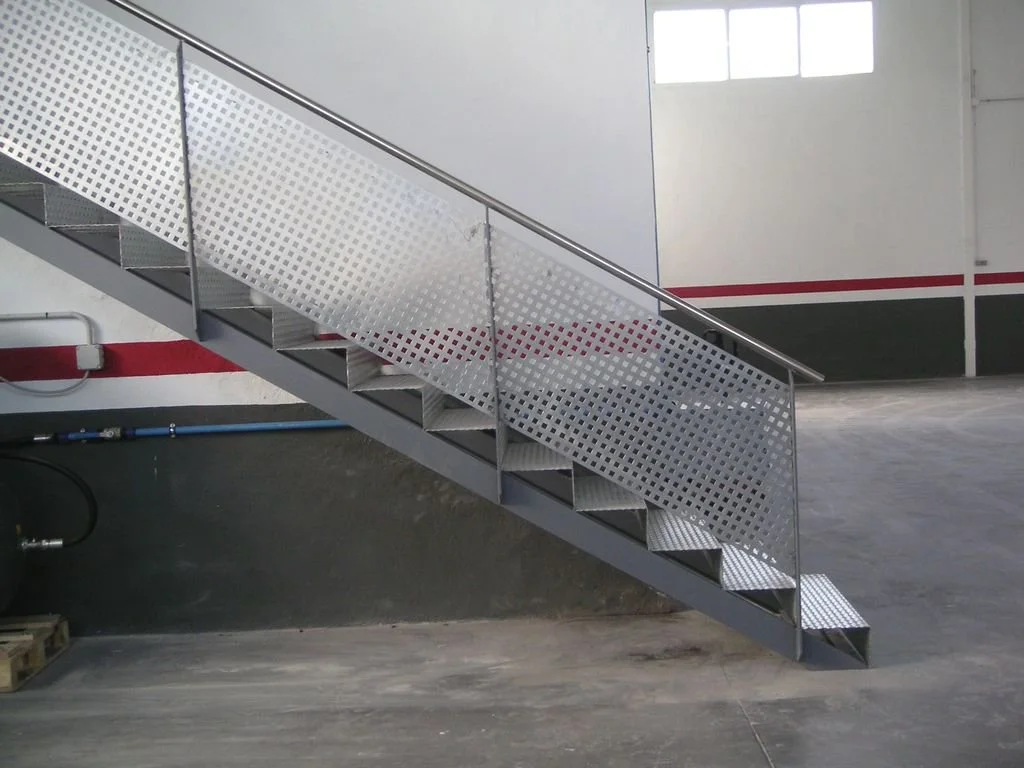 Escalera metálica en un espacio industrial, con barandilla y pasamanos, y paredes blancas y grises.