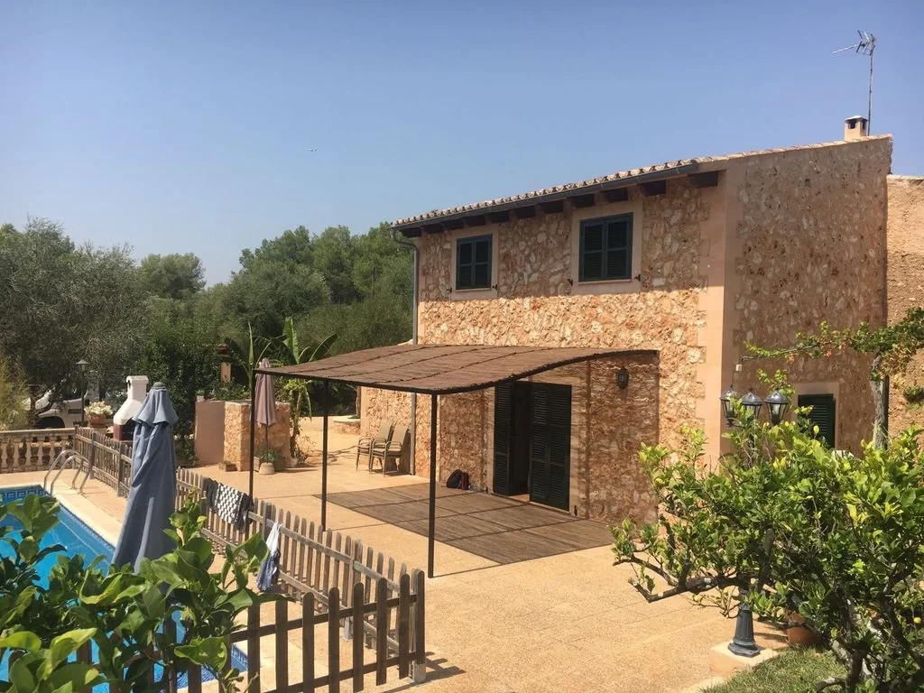 Casa de piedra en un patio con piscina, sombrillas, plantas y árbol con flores.