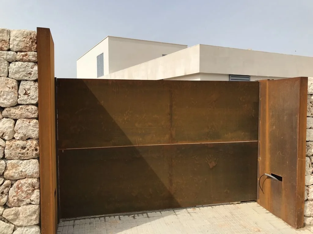 Puerta de metal oxidado en una cerca de piedra, con un muro blanco moderno en el fondo.