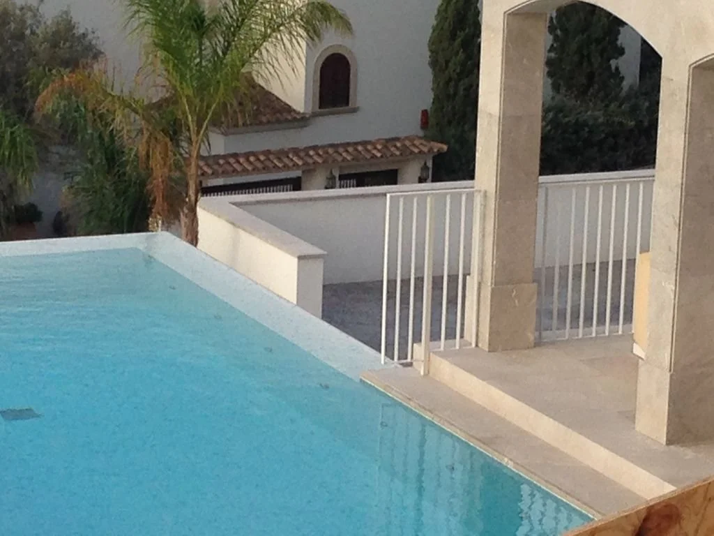 Piscina en un patio con estructura de columnas, rejas blancas y vegetación en el fondo.
