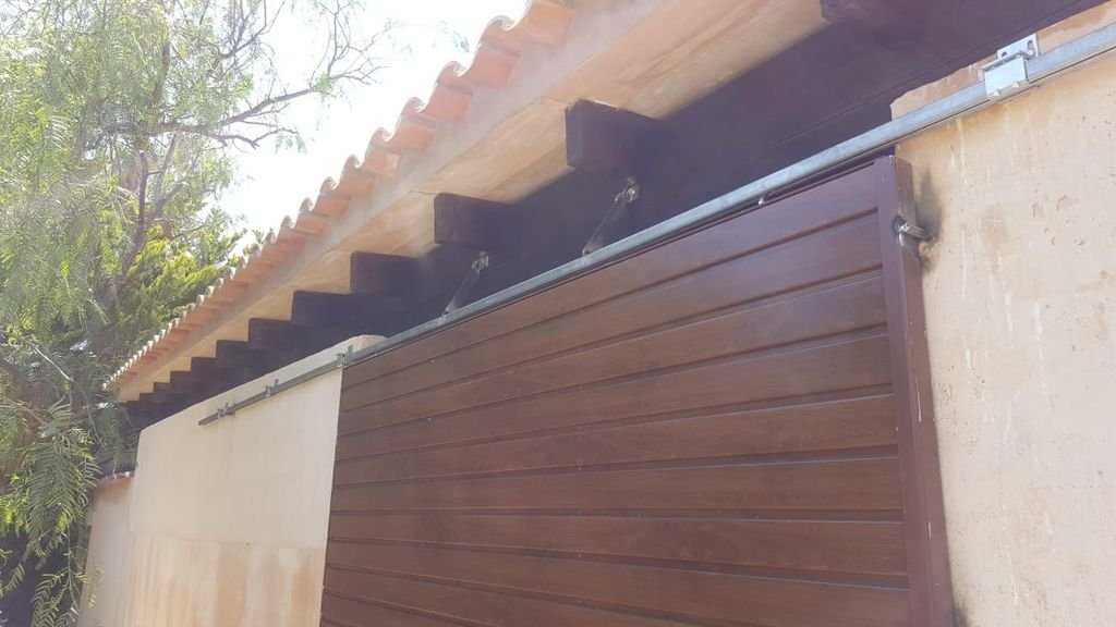 Puerta de garaje de paneles de madera en una pared exterior, con sistema de apertura corredera, bajo un techo con tejas de barro y vigas de soporte.