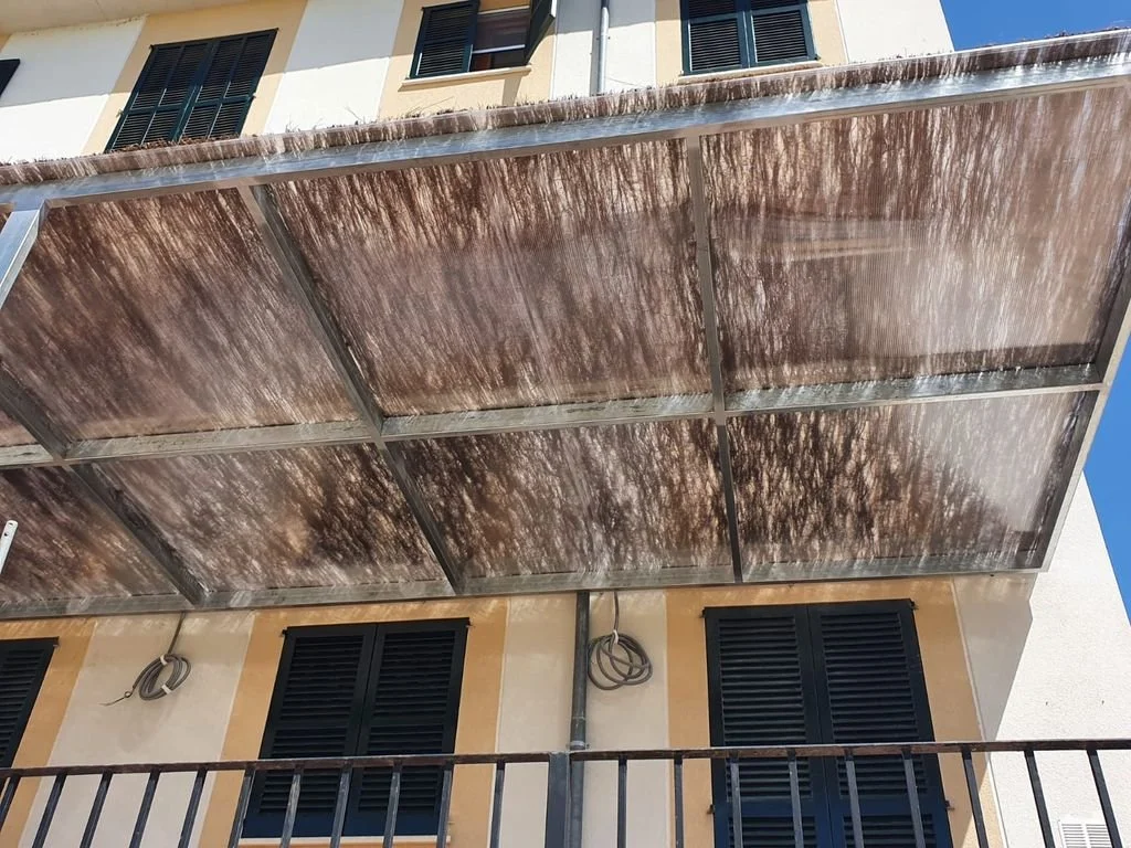 Cubierta de balcón con paneles metálicos oxidados y ventanas con persianas negras en un edificio de madera, con cables eléctricos colgando.