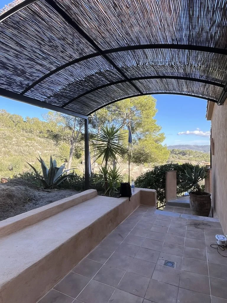 Terraza con toldo de paja, plantas de exterior y vista al campo con árboles y cielo azul.
