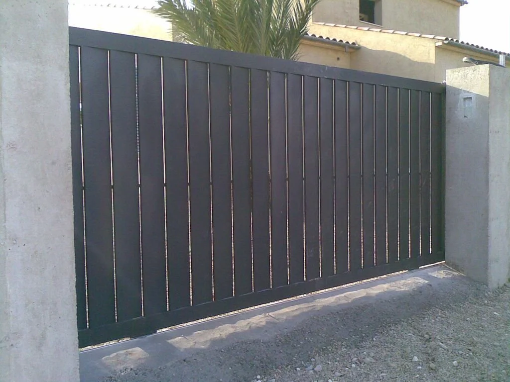 Puerta metálica de color oscuro que cierra una entrada a un área privada, con dos muros de piedra a los lados y plantas en el fondo.
