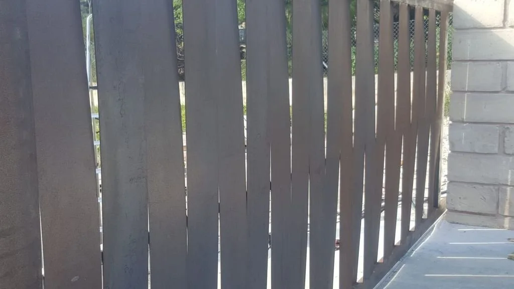 Reja de verja de metal gris con espacio entre barras, junto a muro de ladrillos blancos y una esquina de concreta.