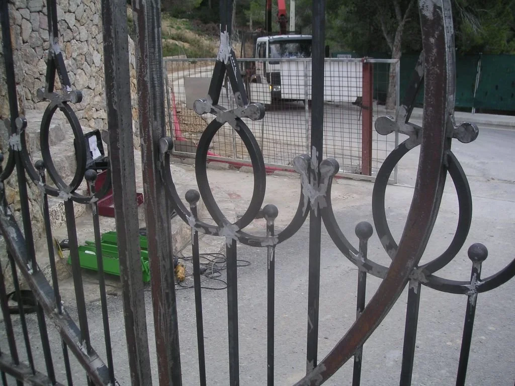 Barra de metal con decoraciones circulares y detalles moldurados, parte de una verja o puerta de hierro. Al fondo, hay una calle, un vehículo blanco, vegetación y un área cerrada con malla y estructura metálica.