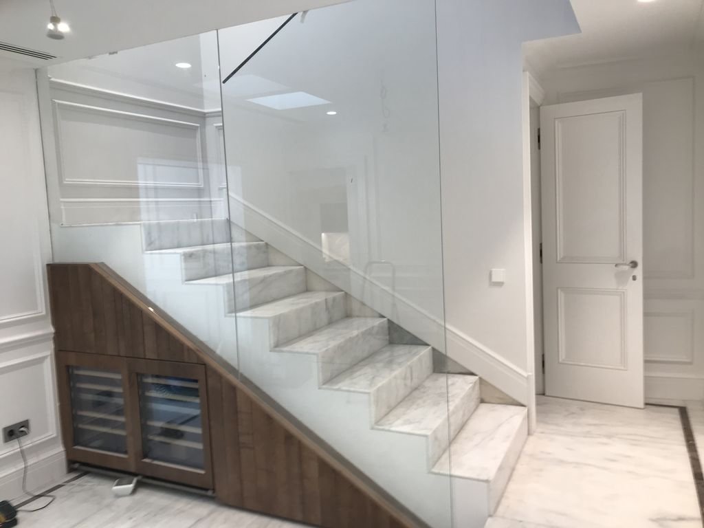 Escalera de mármol con barandilla de cristal y mueble de madera. Ambiente interior con puertas blancas y paredes decoradas con molduras.