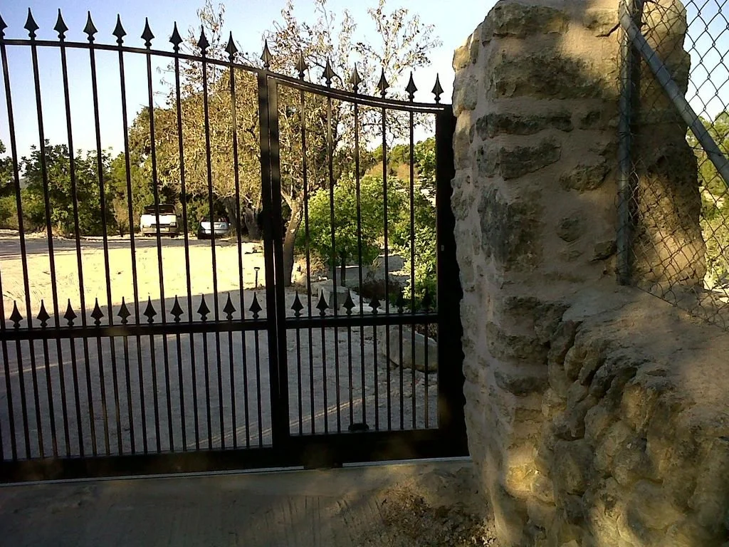 Una puerta de hierro con púas, junto a una pared de piedra, da acceso a un área con árboles y vehículos estacionados