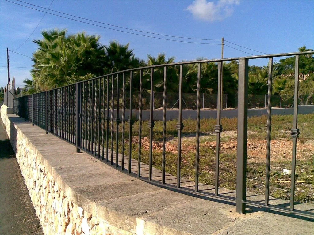 Reja negra decorativa en la parte superior de una cerca de piedra, con árboles y cables eléctricos en el fondo, en un día soleado.